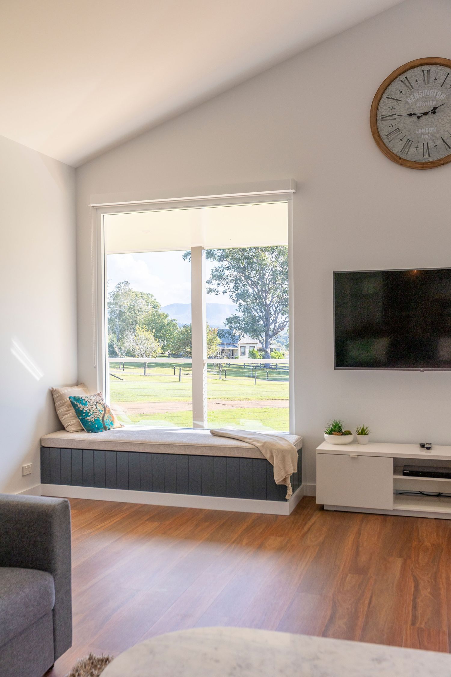 Modern living room with hardwood floors, gray couch, wall-mounted TV, and large windows overlooking a green landscape.