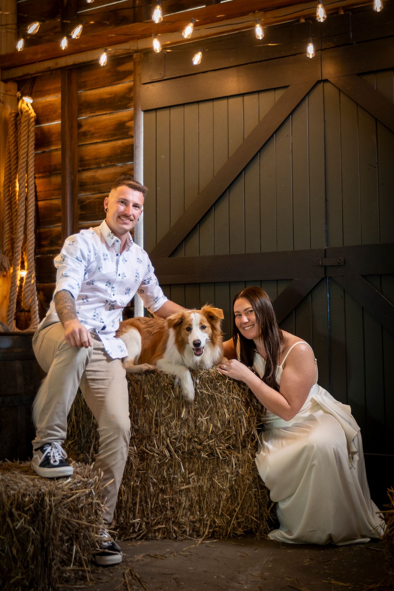 Photo link to the type of photography sessions. Couple sitting on hay bales with their dog inside a rustic barn lit by string lights.