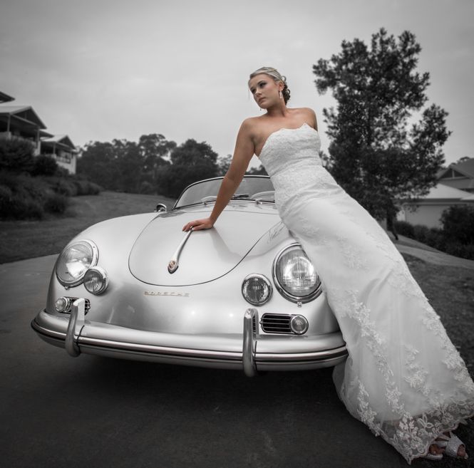 A figure in a white wedding dress leans against a classic silver Porsche 356 sports car on an overcast day.