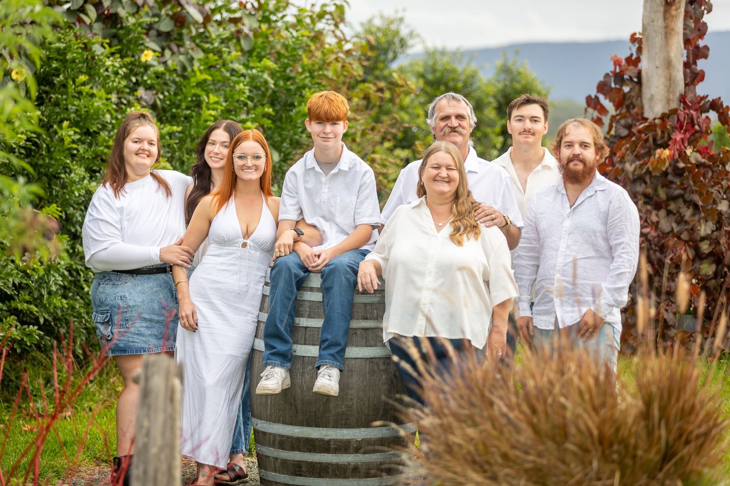 Family group photo outdoors near a wine barrel, wearing white outfits, surrounded by lush greenery.