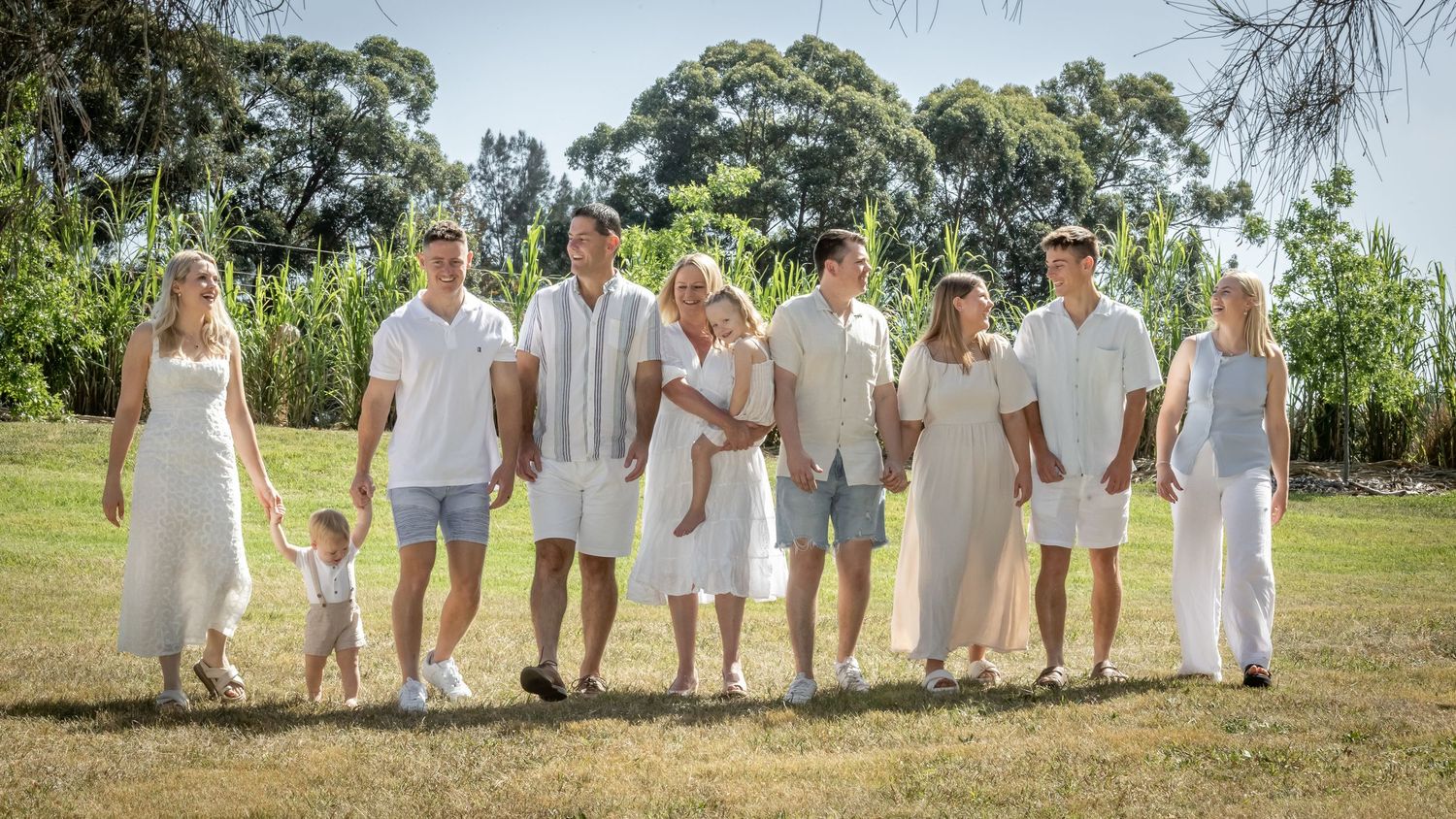Large family group in matching white outfits walking together on green grass with lush trees in background.