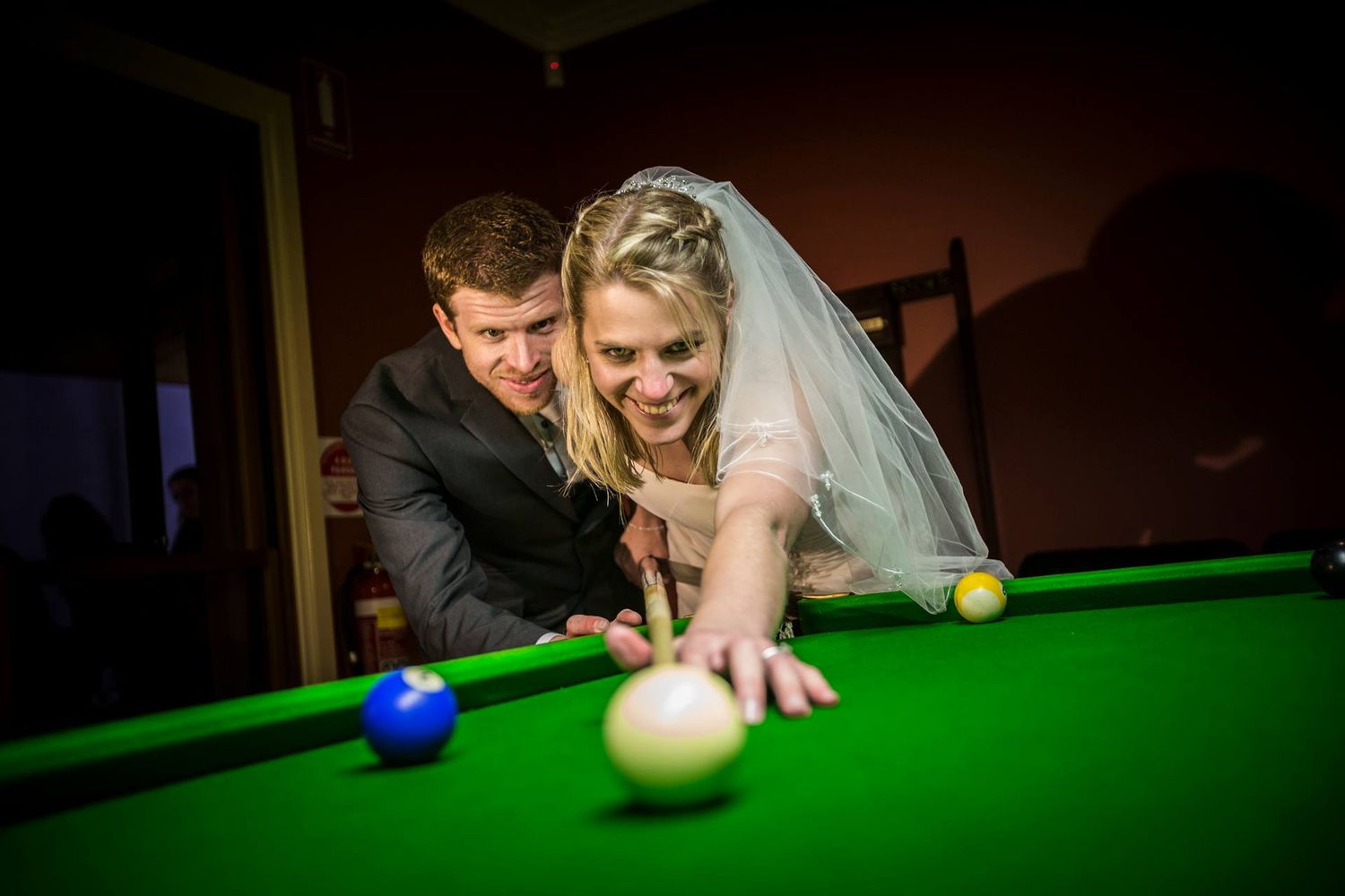 Wedding couple plays snooker together in a dimly lit room with unique action shots of them taking their shots at the table.