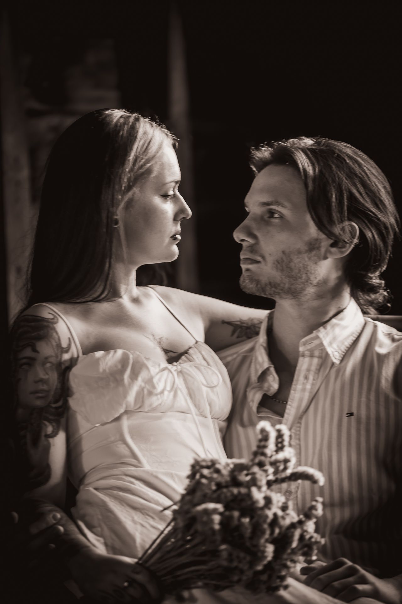 Black and white photo of a tattooed bride in white dress gazing at groom holding flowers.