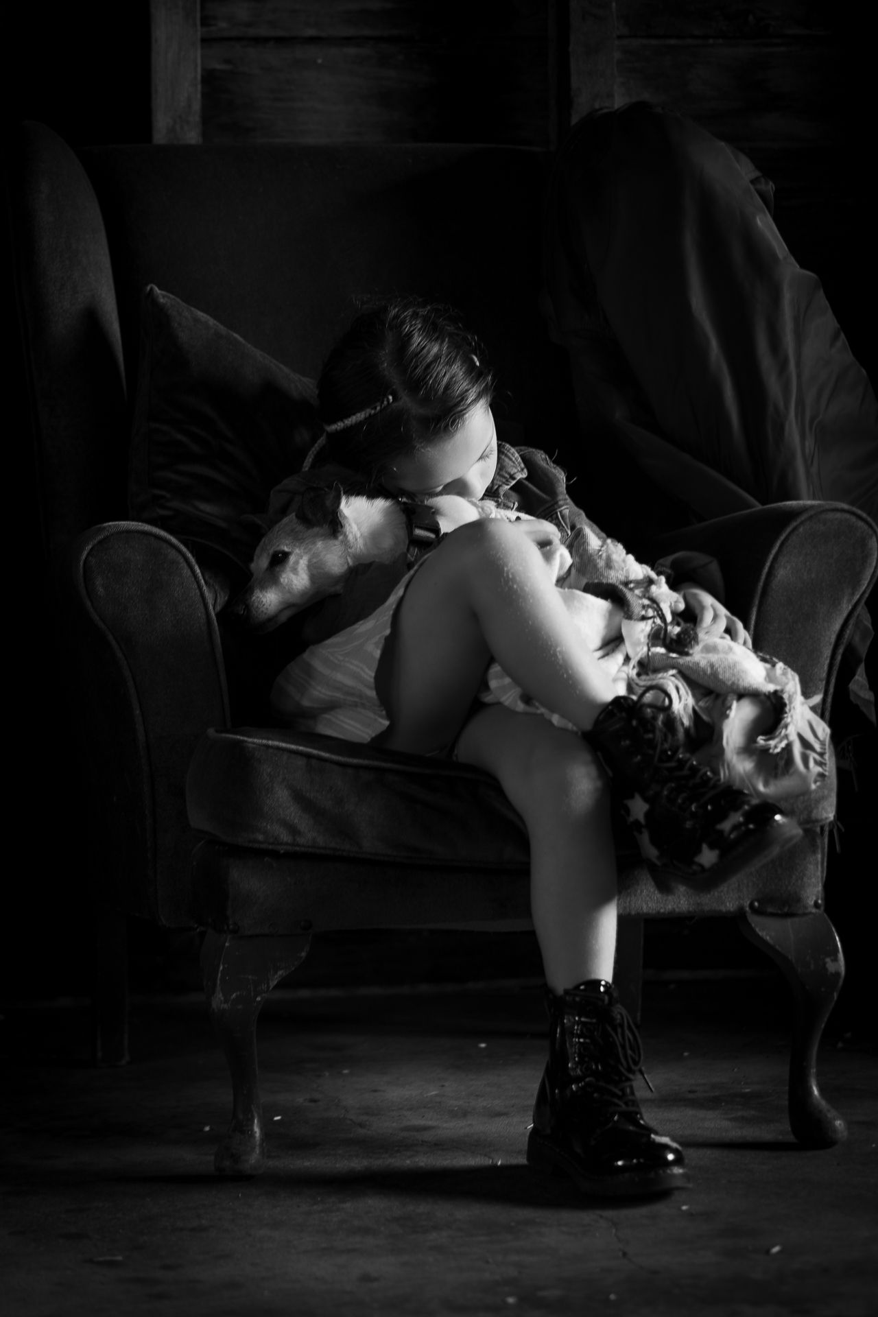 Black and white photo of a young woman curled up in a vintage armchair, wearing boots, in a moody dark setting.