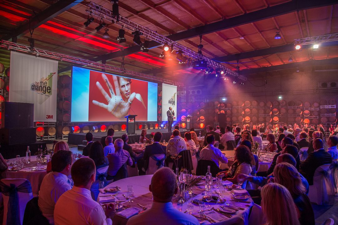 Large banquet hall with round tables and guests during an event with red and blue lighting and projector screens.