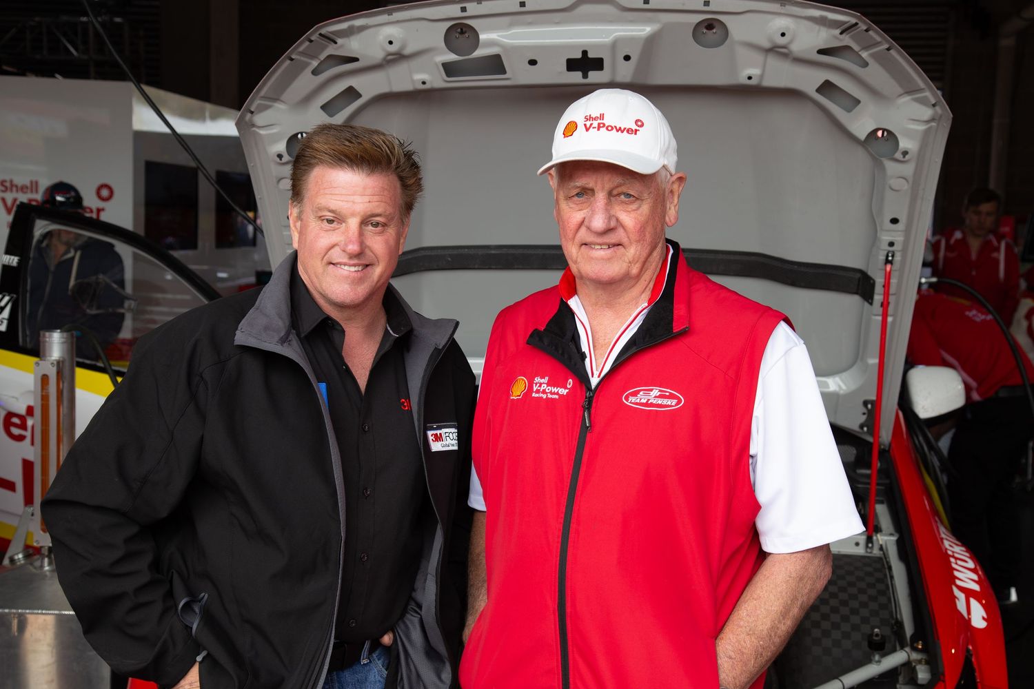 Racing team members in red and black uniforms gather around equipment in a professional motorsports pit area. Chip Foose and Dick Johnson