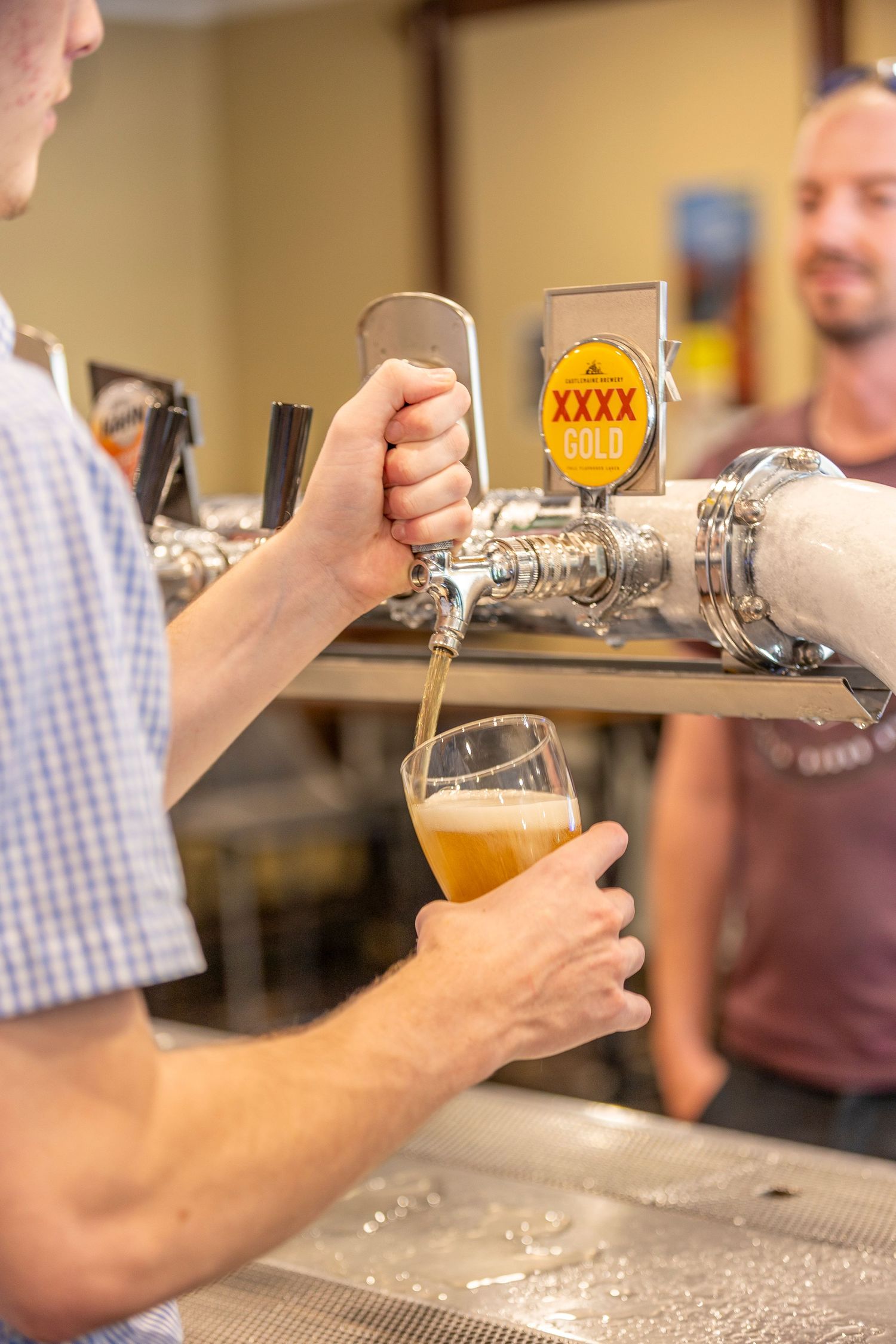 A bartender pours a draft beer from a tap with a XXXX logo into a glass at the bar counter.