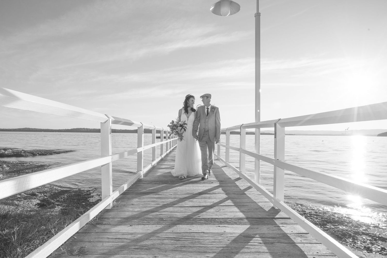 A couple stands together on a wooden pier overlooking the ocean at sunset in black and white photography.