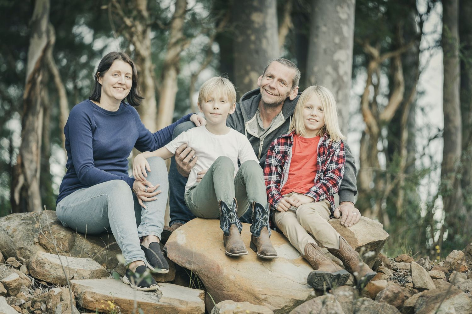 A group sits together on a large rock in a forest setting wearing casual outdoor clothing.