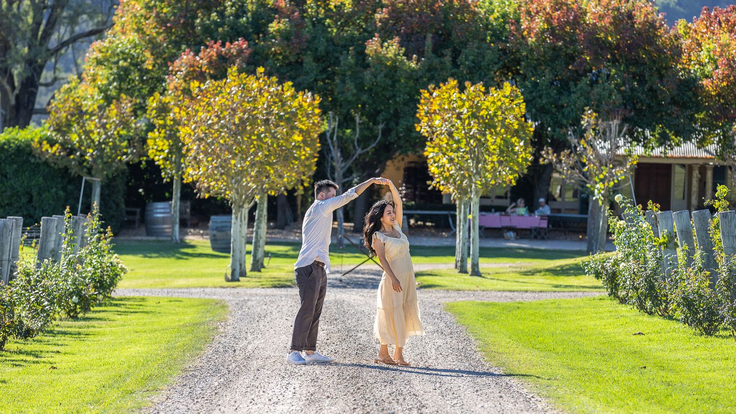 Couple dancing on a tree-lined gravel path in a lush green garden on a sunny day.