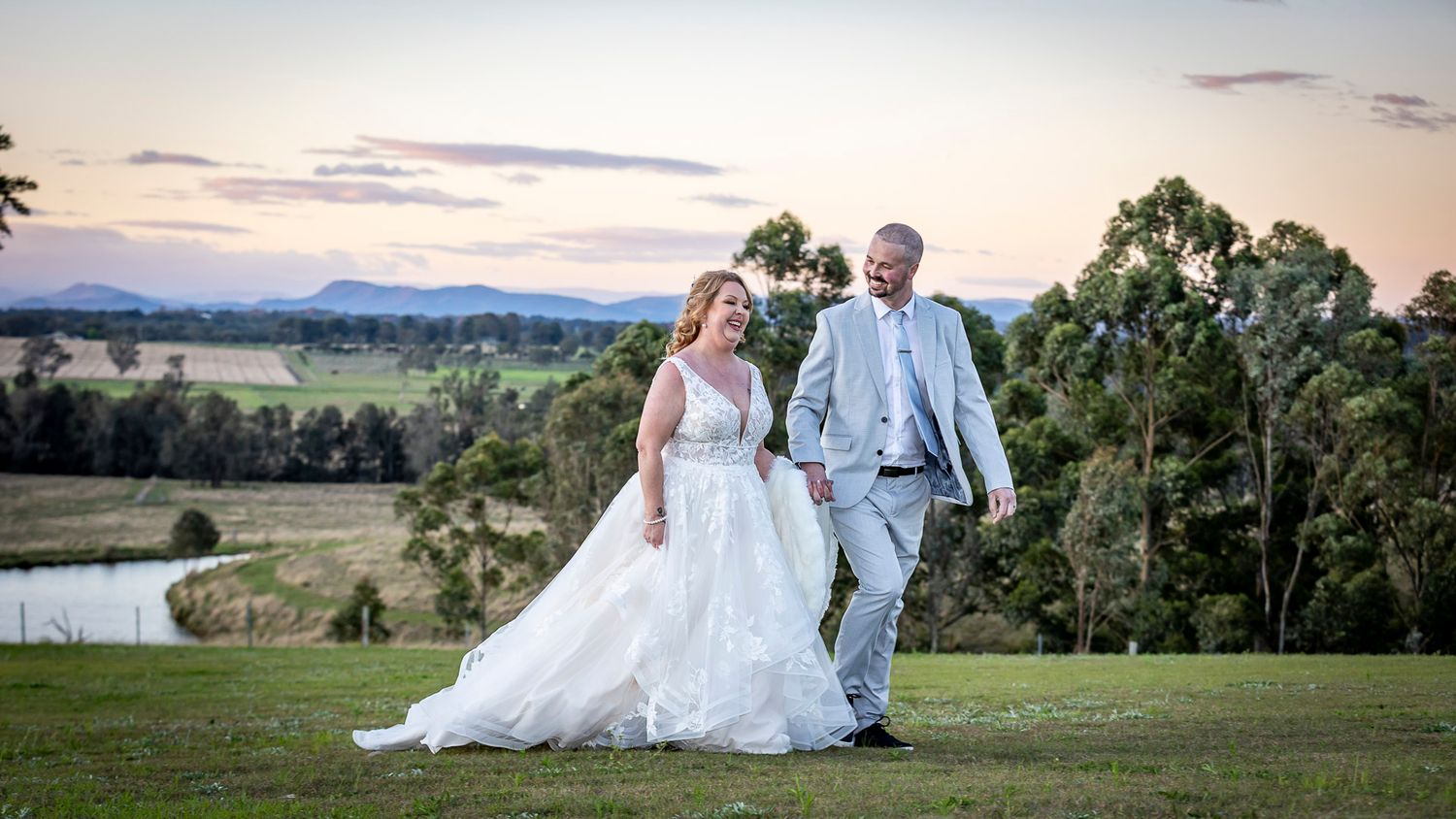 Bride and groom walking hand in hand on a lush green hill at sunset with scenic countryside views.