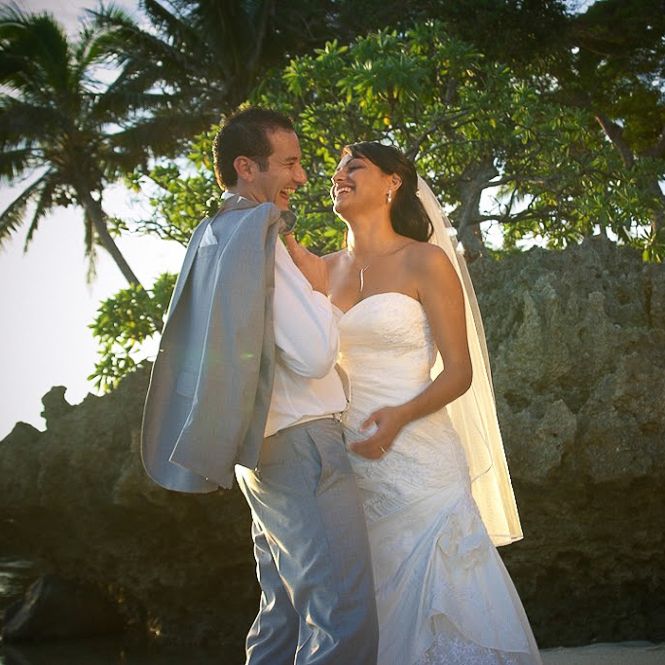 Couple shares tender moment at tropical beach wedding with palm trees and natural stone backdrop at sunset.