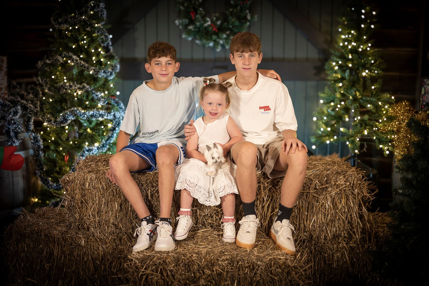 Three people sit on hay bales in front of a decorated Christmas tree with twinkling lights in the background.
