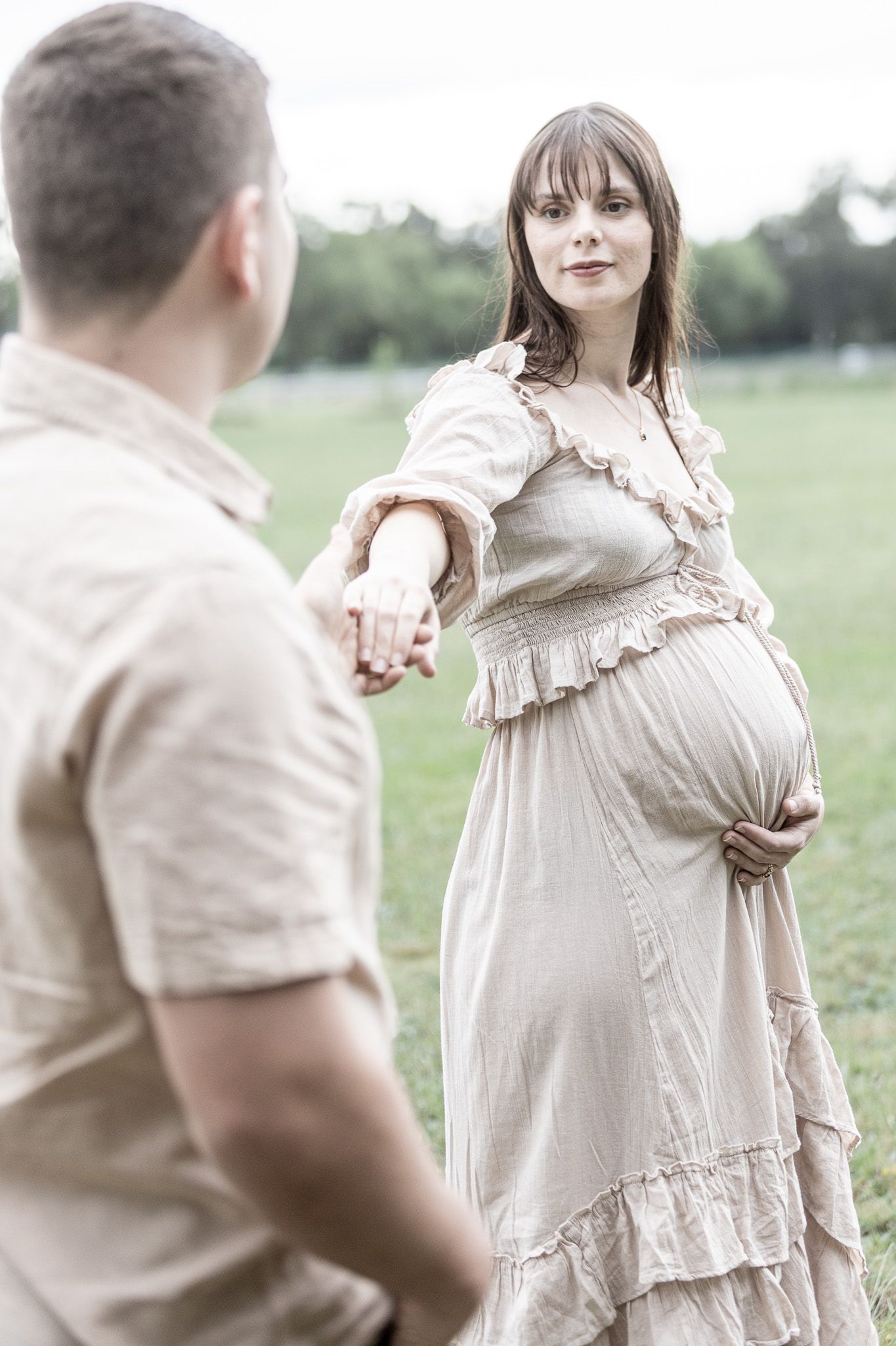 A romantic couple in light beige clothing poses together in a dreamy outdoor field setting for a maternity photos
