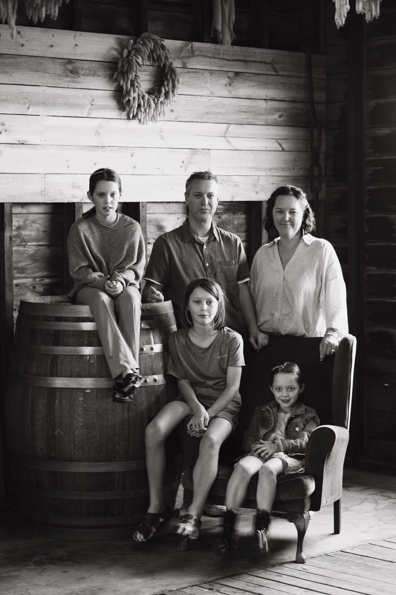 Black and white family portrait of five people posing in a rustic barn with a barrel and vintage chair.