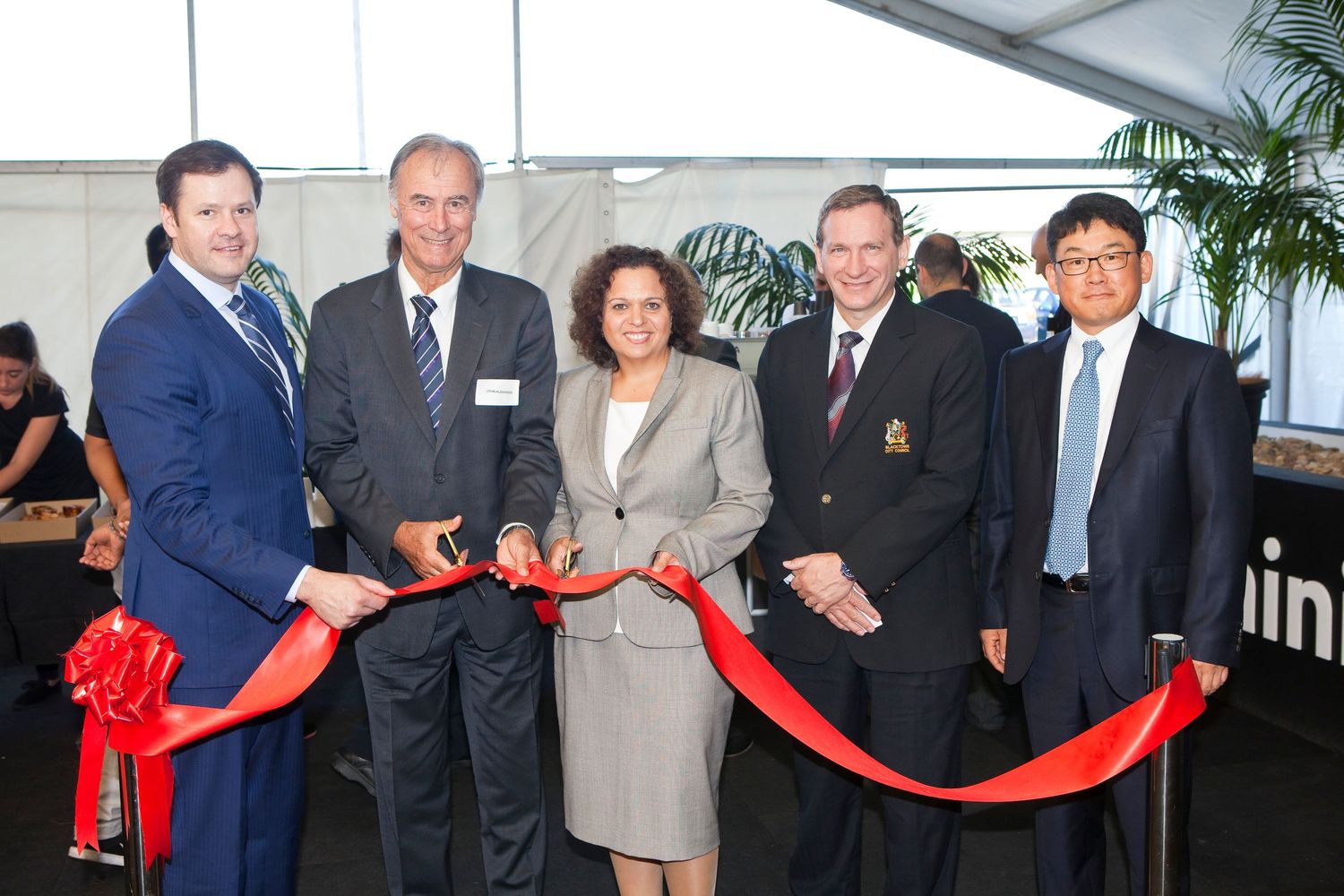 Group of people in business attire gathered at a ceremonial red ribbon cutting event in a bright indoor space.
