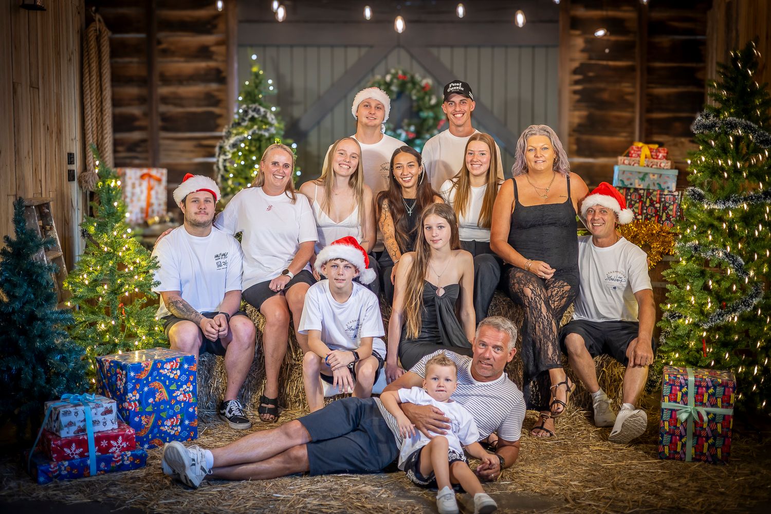 Large family Christmas photo in rustic barn with decorated trees, Santa hats, and wrapped gifts.