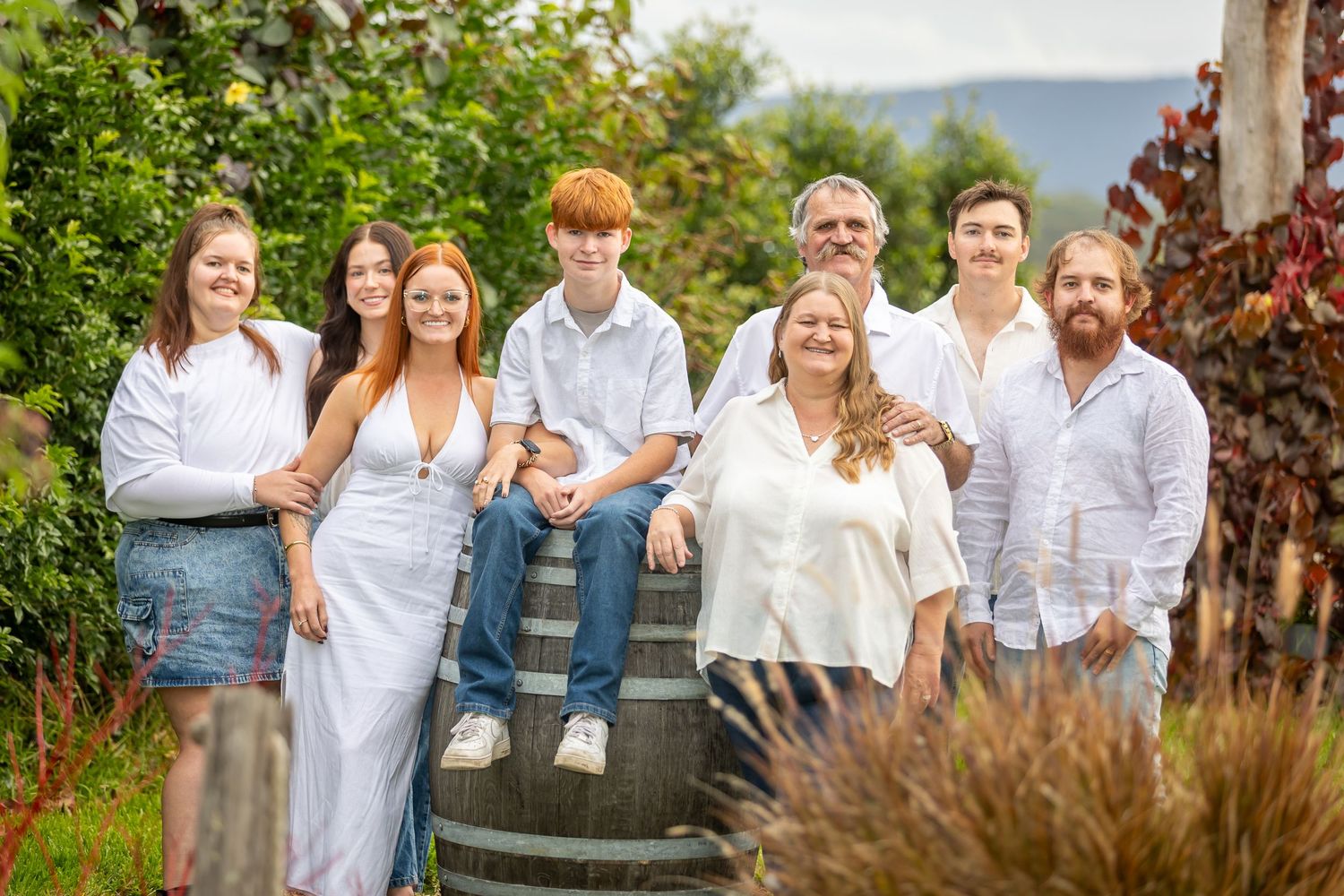 Large family group portrait outdoors in vineyard setting, wearing coordinated white outfits, smiling together on sunny day.