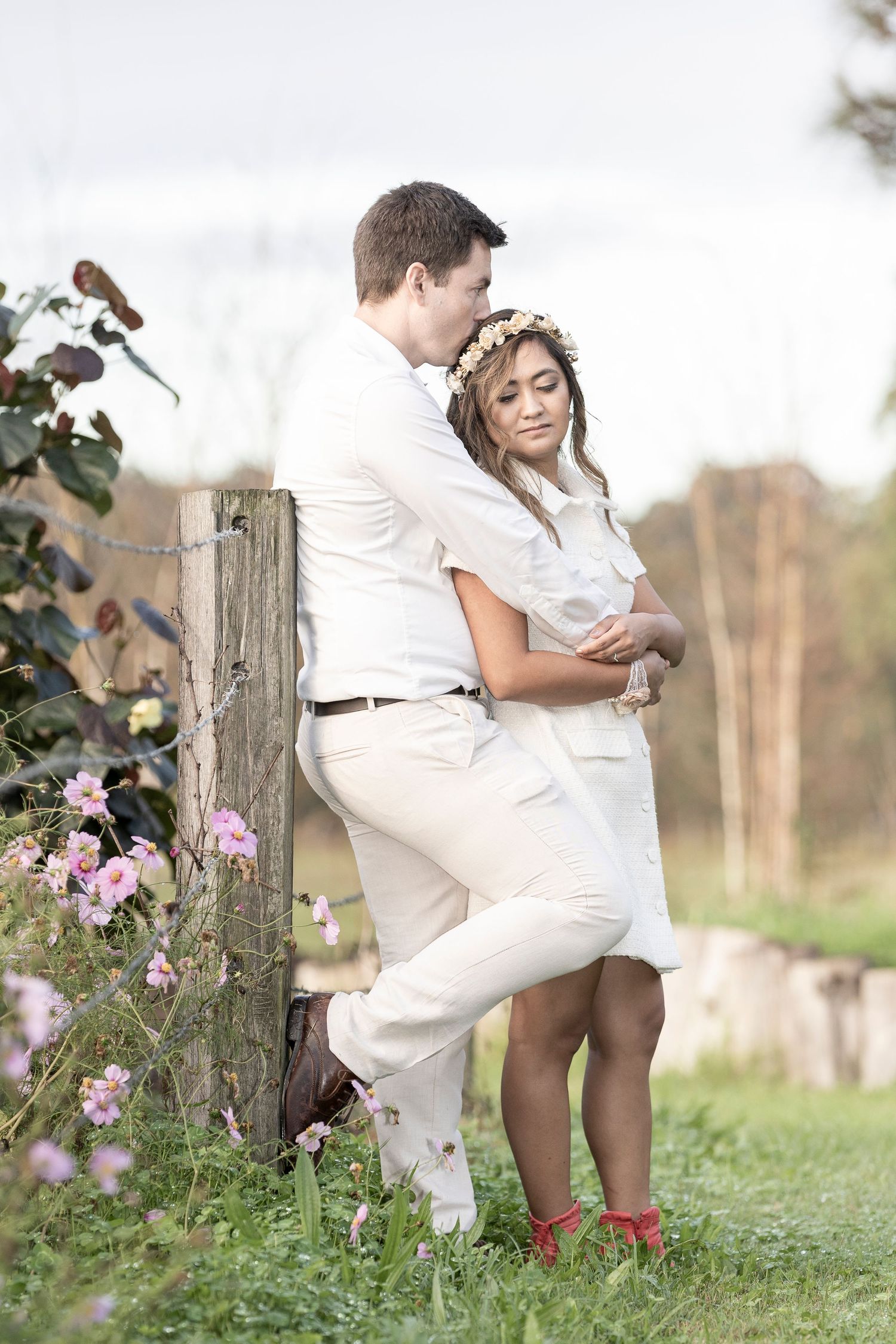 A romantic couple in white outfits poses together on a rustic fence post surrounded by delicate pink wildflowers.