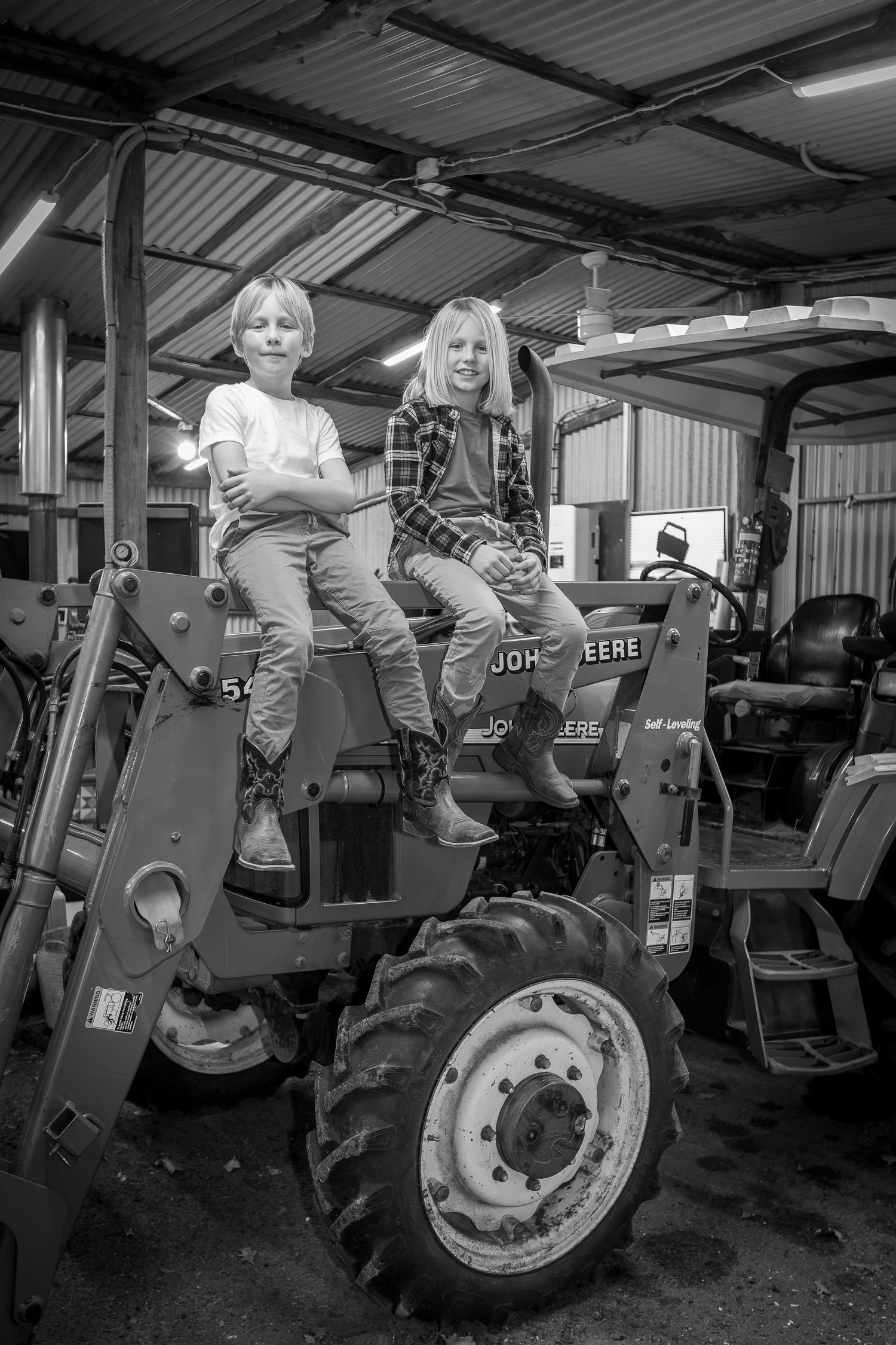 Two children pose atop a parked tractor in a black and white farm setting.