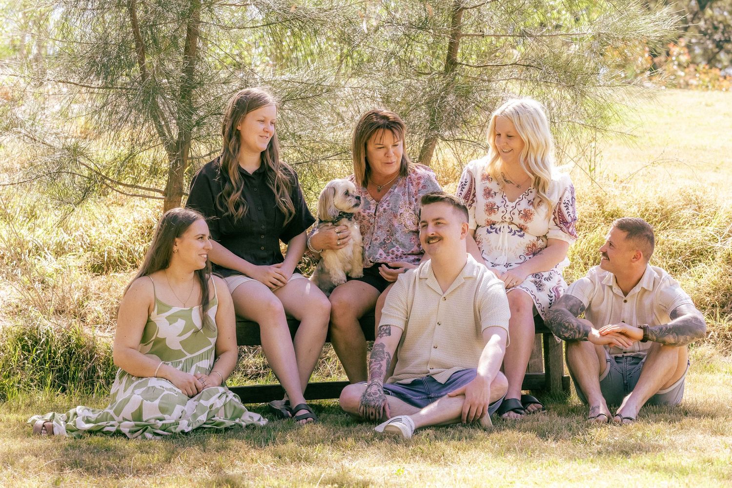 Family group photo with dog sitting on grass outdoors among trees on a sunny day.