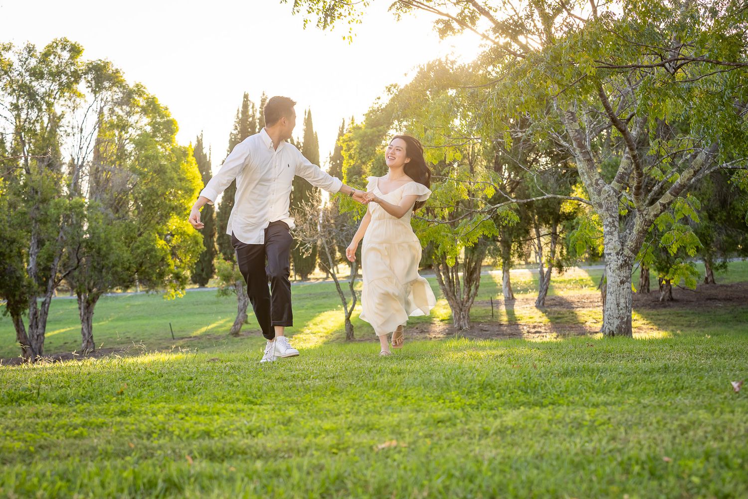 A joyful couple in wedding attire runs hand-in-hand through a sunlit green park surrounded by trees.