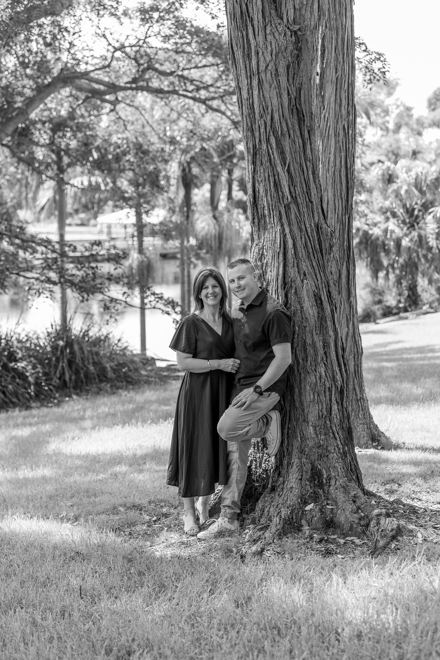 Romantic black and white portrait of a couple posing together beneath a large tree trunk in a serene park setting.