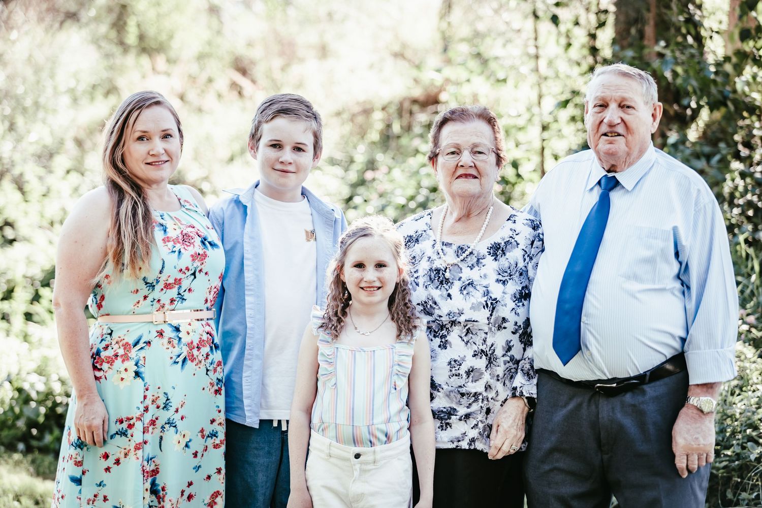 Family of five posing outdoors, including two children and grandparents, dressed in smart casual attire amid lush greenery.