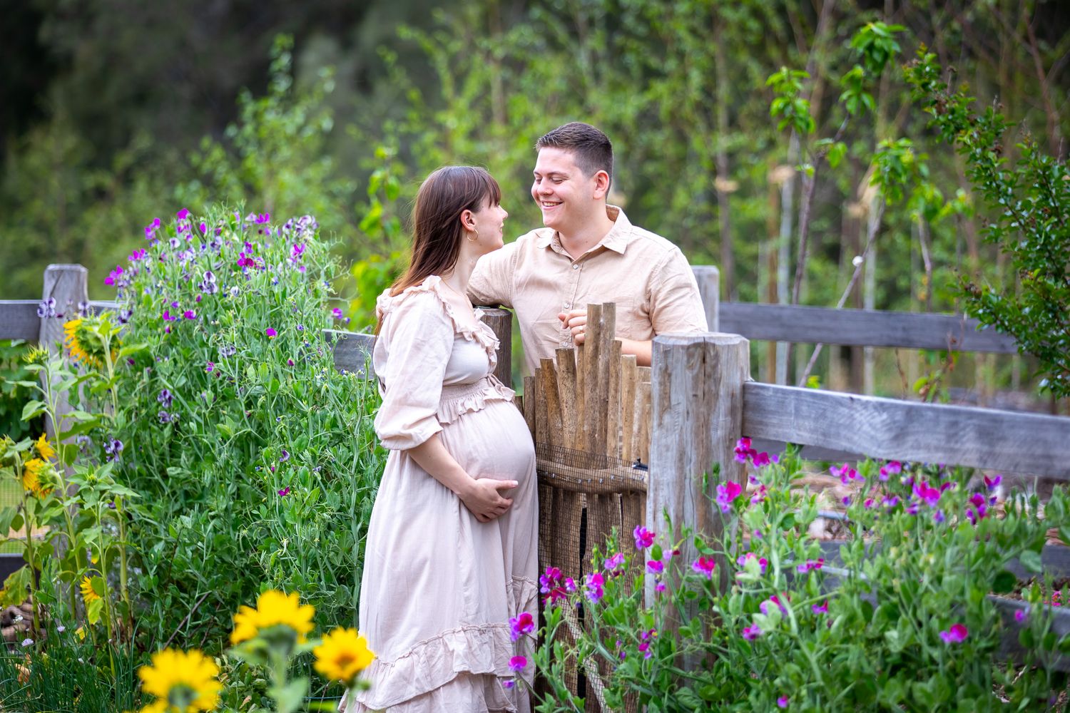 Couple sharing romantic moments in a lush garden setting with colourful flowers and rustic wooden fence for a maternity photos