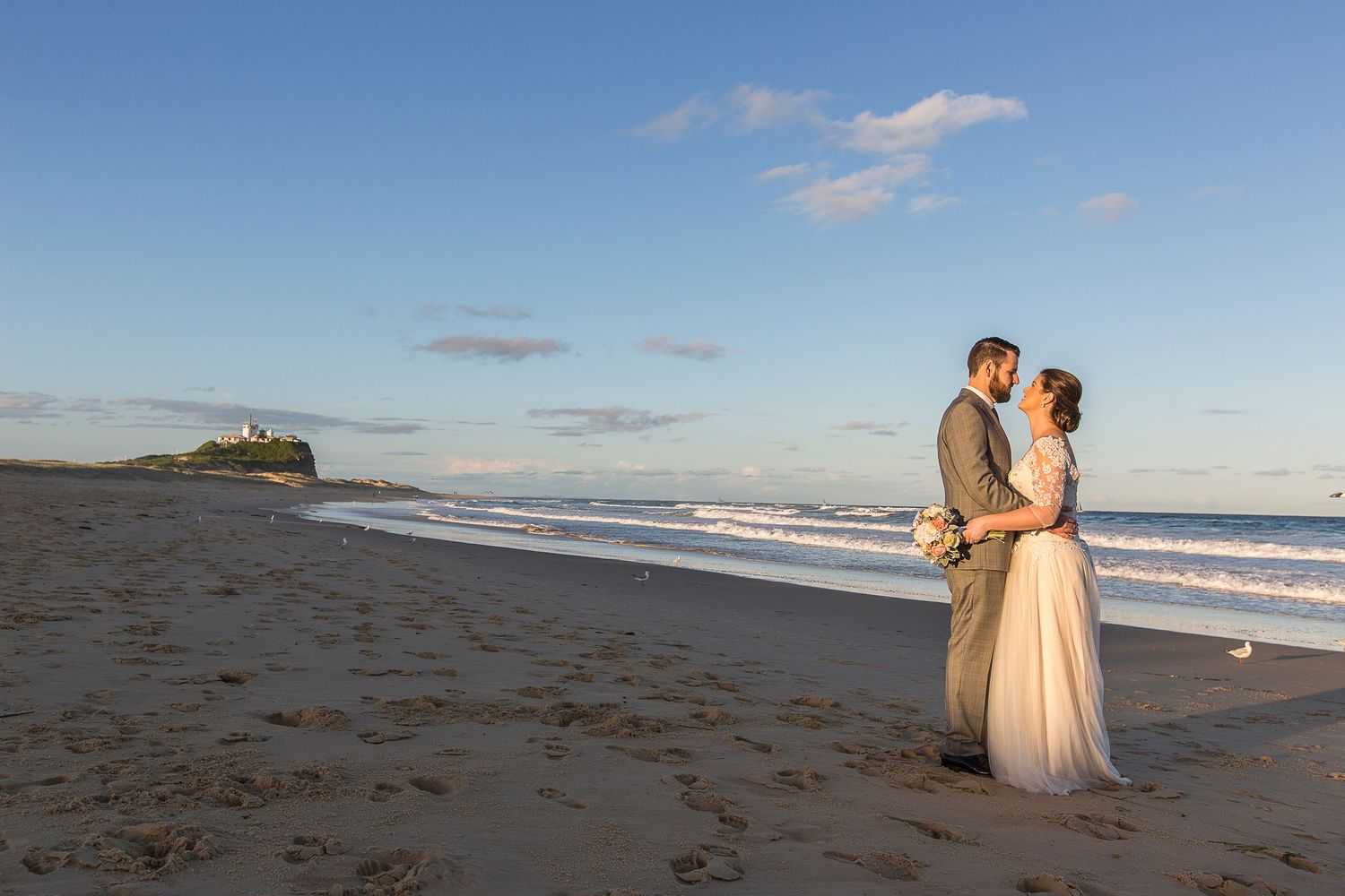Two people embrace on a sandy beach at sunset with Nobska Lighthouse visible on a hill in the background.