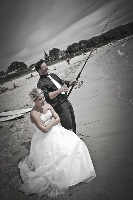 A wedding couple poses on a beach with the groom holding a fishing rod while the bride sits in her white dress.