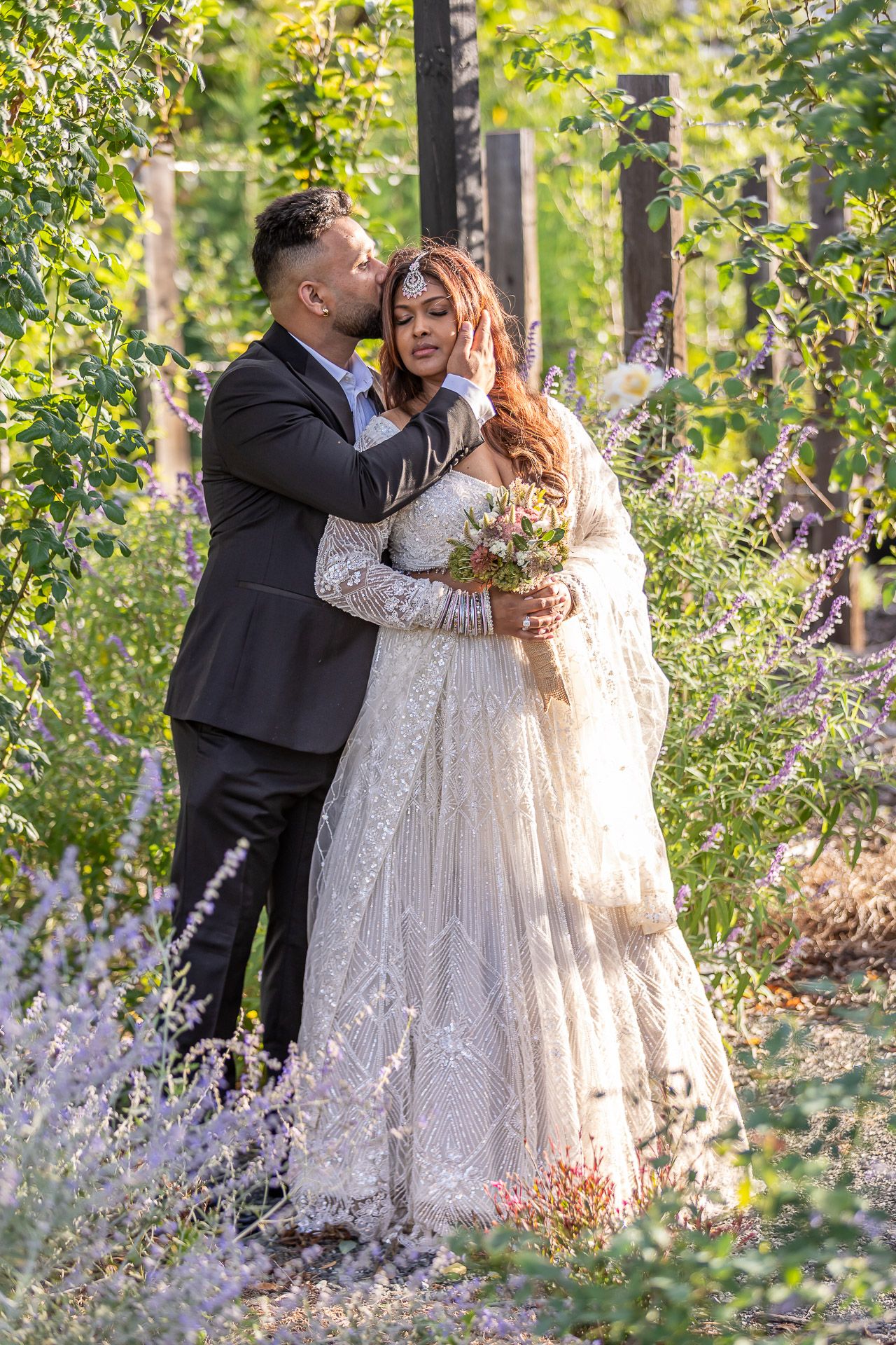 A romantic wedding couple embraces in a garden filled with lavender and greenery on a sunny afternoon.