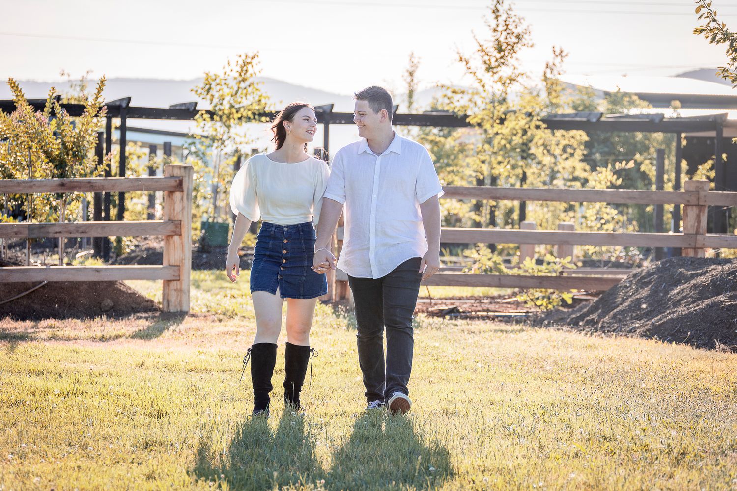 A couple holds hands while walking through a sunlit autumn field during golden hour.