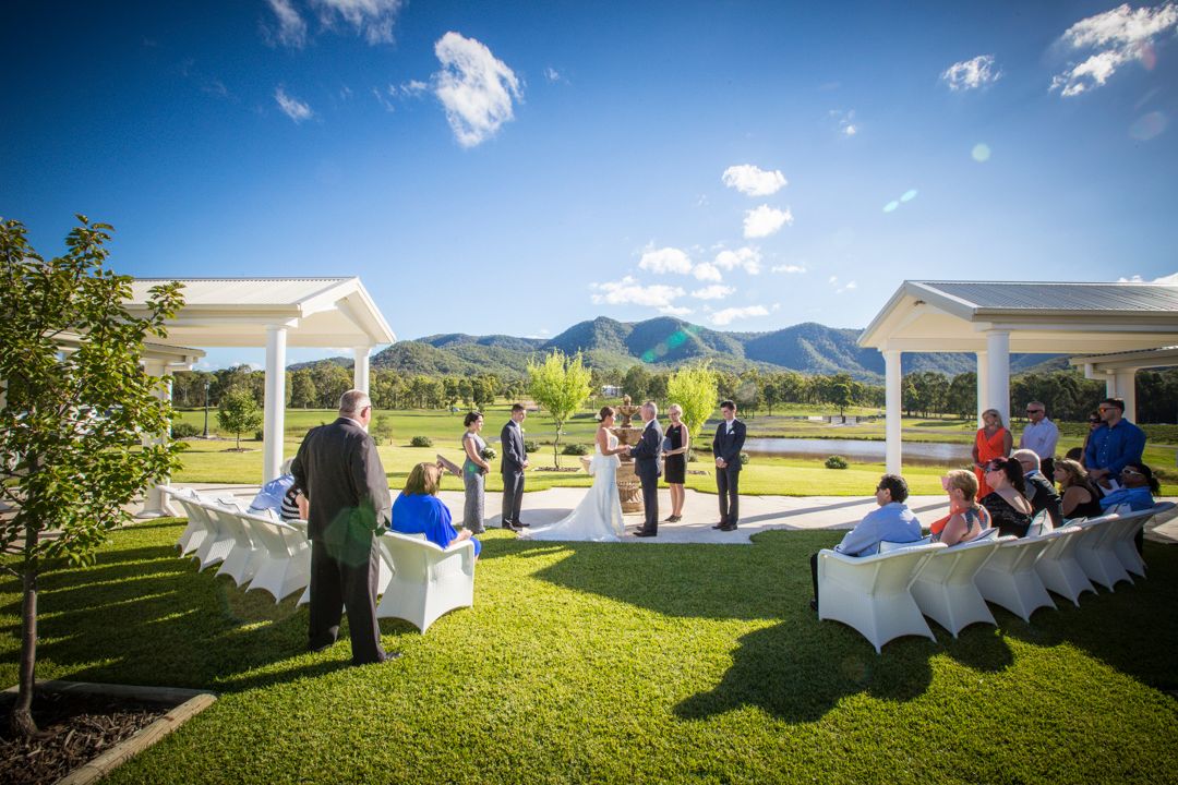 Outdoor wedding ceremony setup on a green lawn with white chairs and a decorative arch against a bright blue sky.