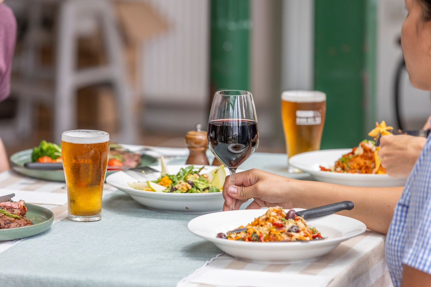 Dinner table with plates of food, glasses of red wine and beer being enjoyed by diners at a casual restaurant setting.