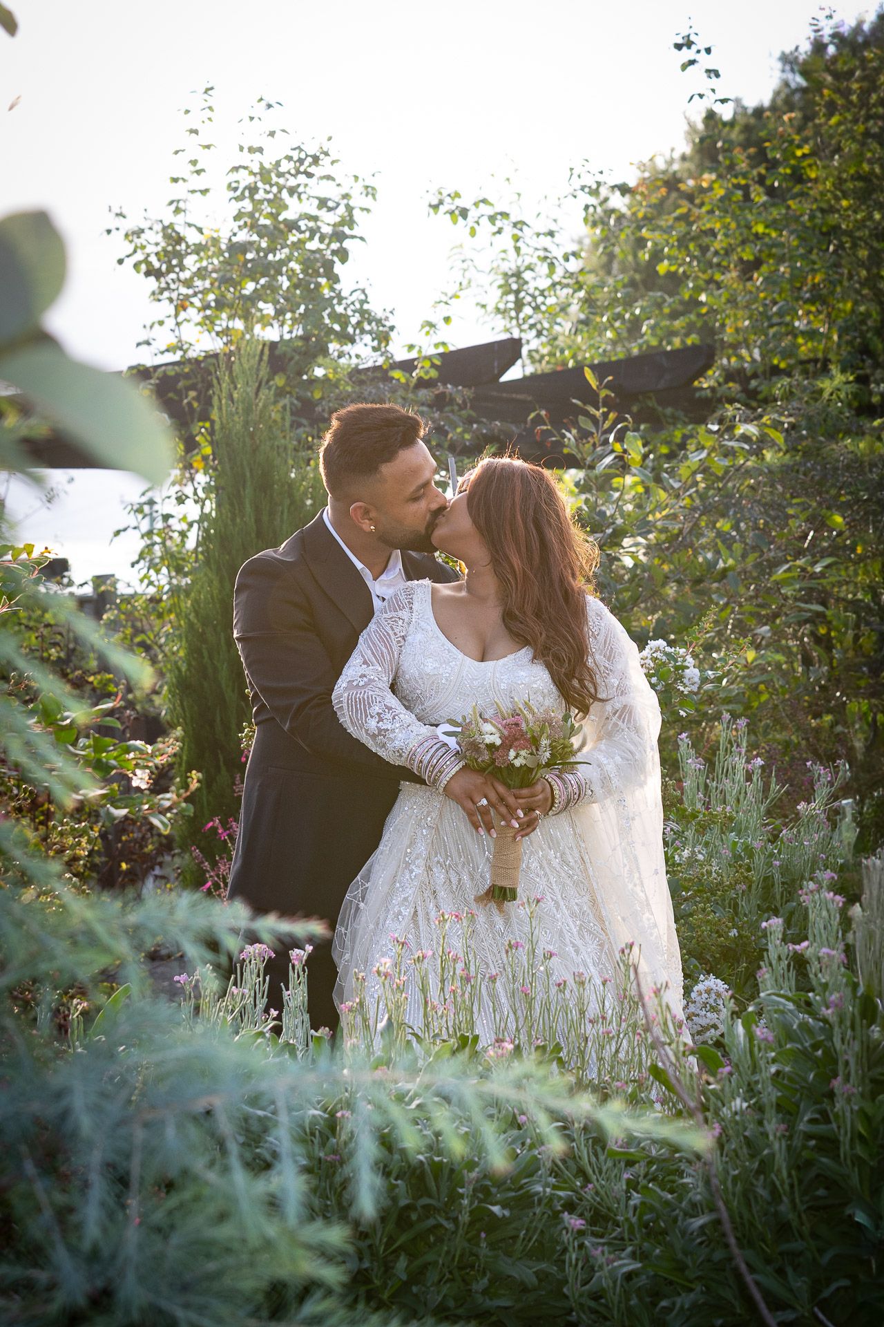A romantic couple in white lace embraces in a lush garden setting during golden hour.