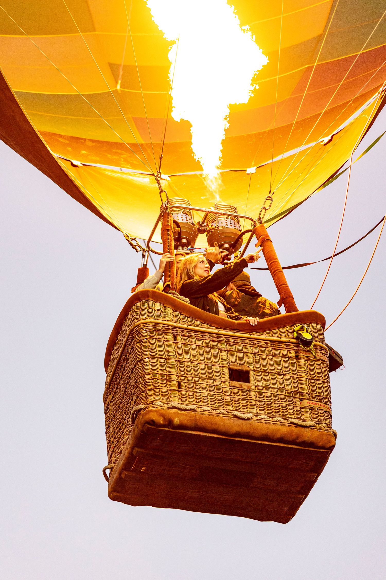 A hot air balloon basket illuminated by glowing burners against a bright orange sky during flight.