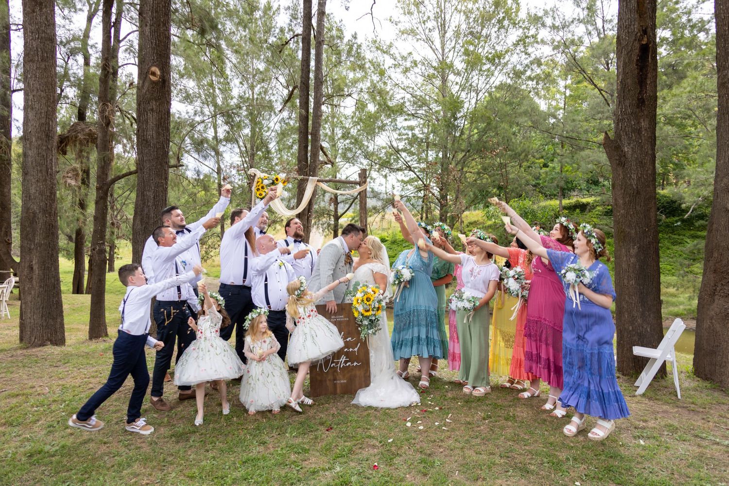 Large group in colorful attire dancing and celebrating together among trees in an outdoor woodland setting.