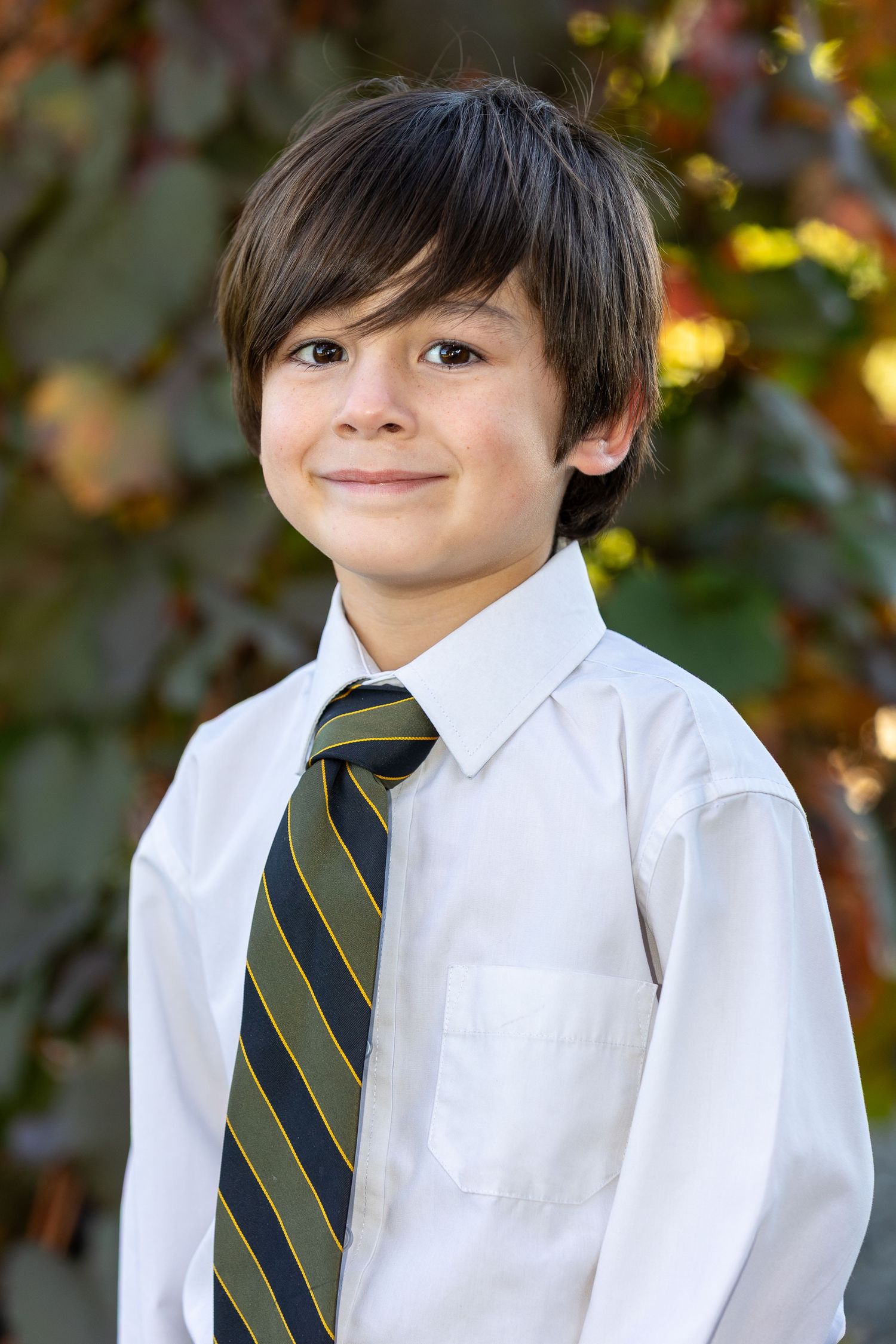Young boy in white dress shirt and striped tie smiling outdoors with colorful autumn foliage background.