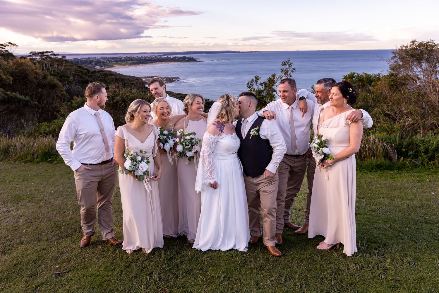Wedding party in light colored formal attire poses together on scenic coastal hilltop at dusk.