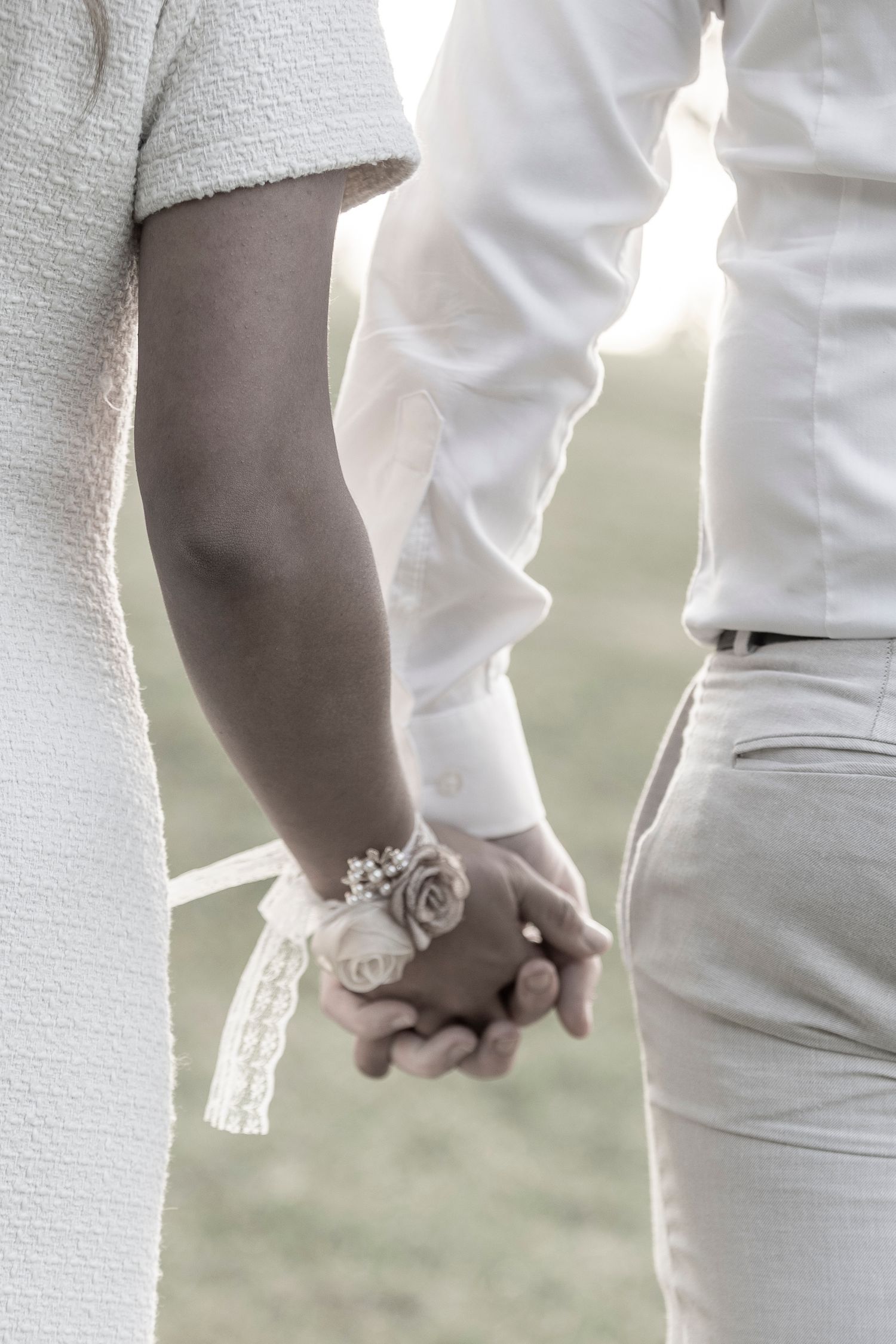 A couple holds hands intimately while wearing white attire, with an elegant rose corsage adorning their clasped hands.