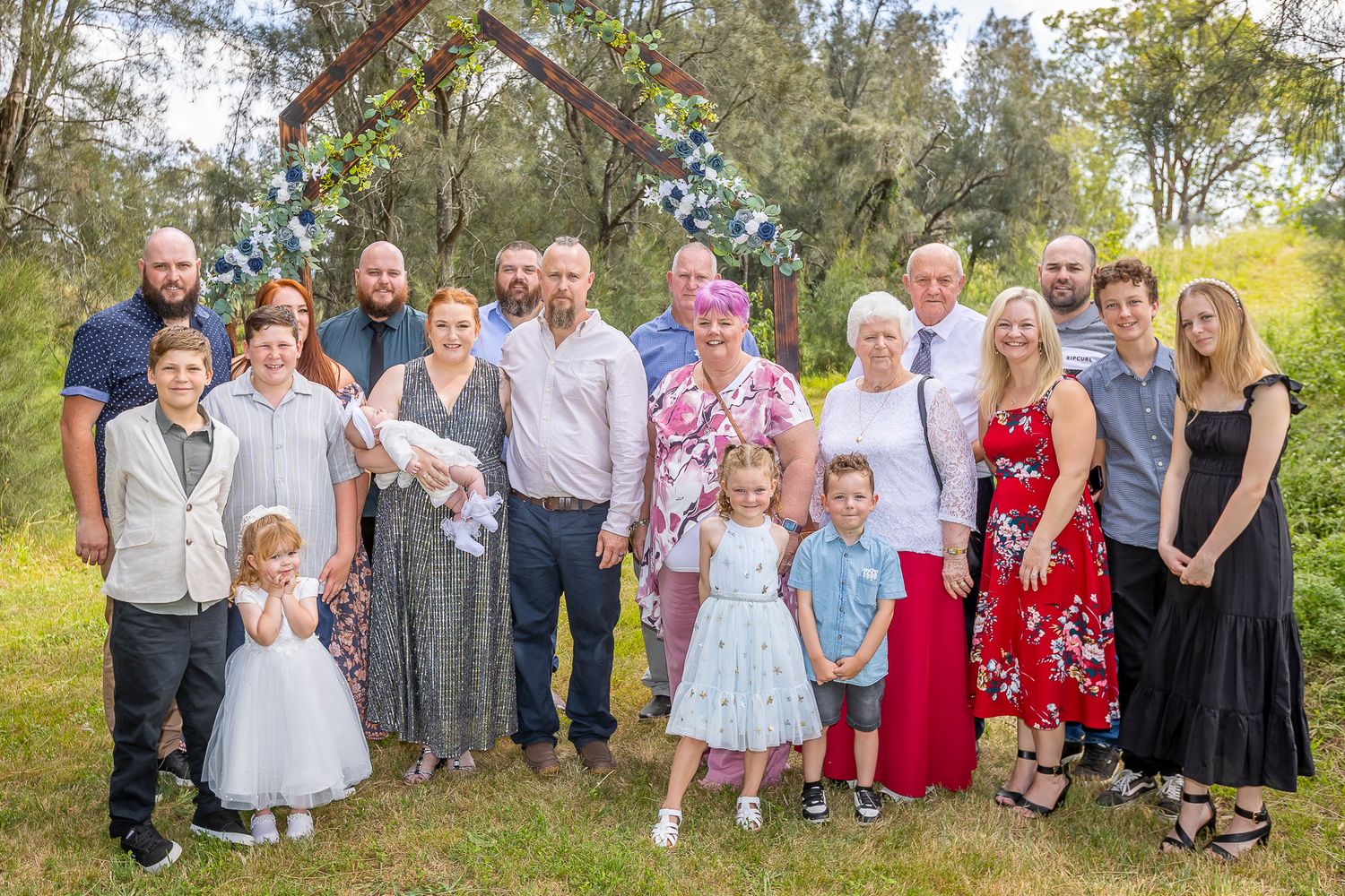 Large group of wedding guests posing together outdoors in a garden setting with trees in the background.
