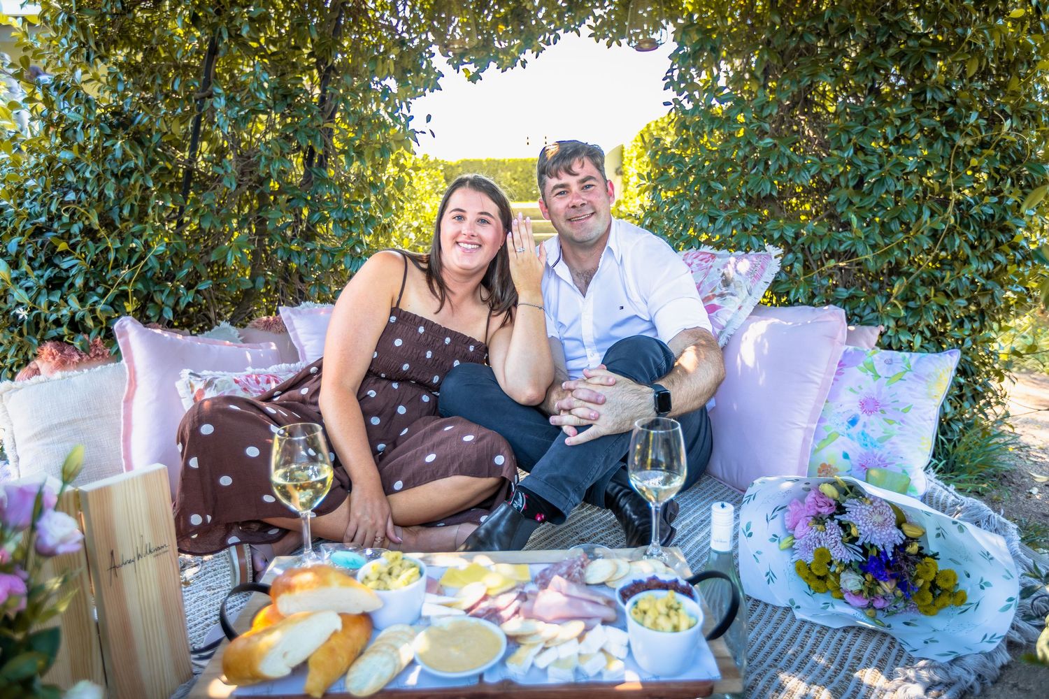 Couple smiling on outdoor picnic setup with wine glasses, cheese board, and lush green hedges in background.