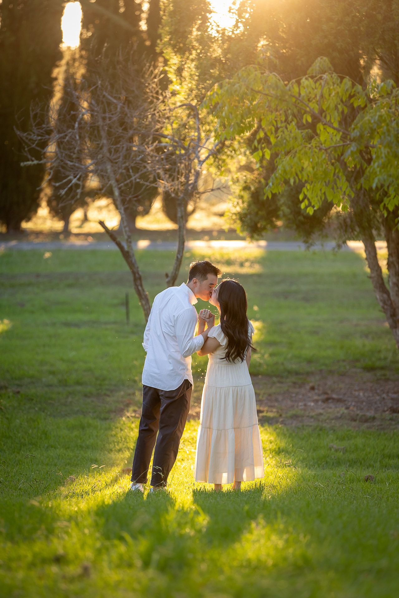 Couple sharing a romantic moment at golden hour in a lush green park, woman in white dress holding a fan.