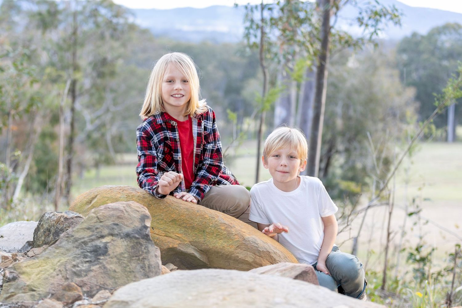 Two children sit on a large rock formation with mountains and trees in the background.