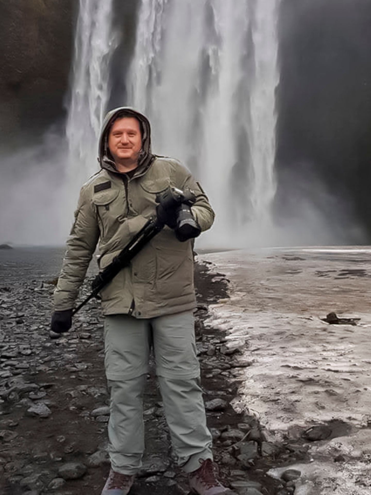 Person wearing outdoor gear standing near a powerful waterfall on rocky terrain.