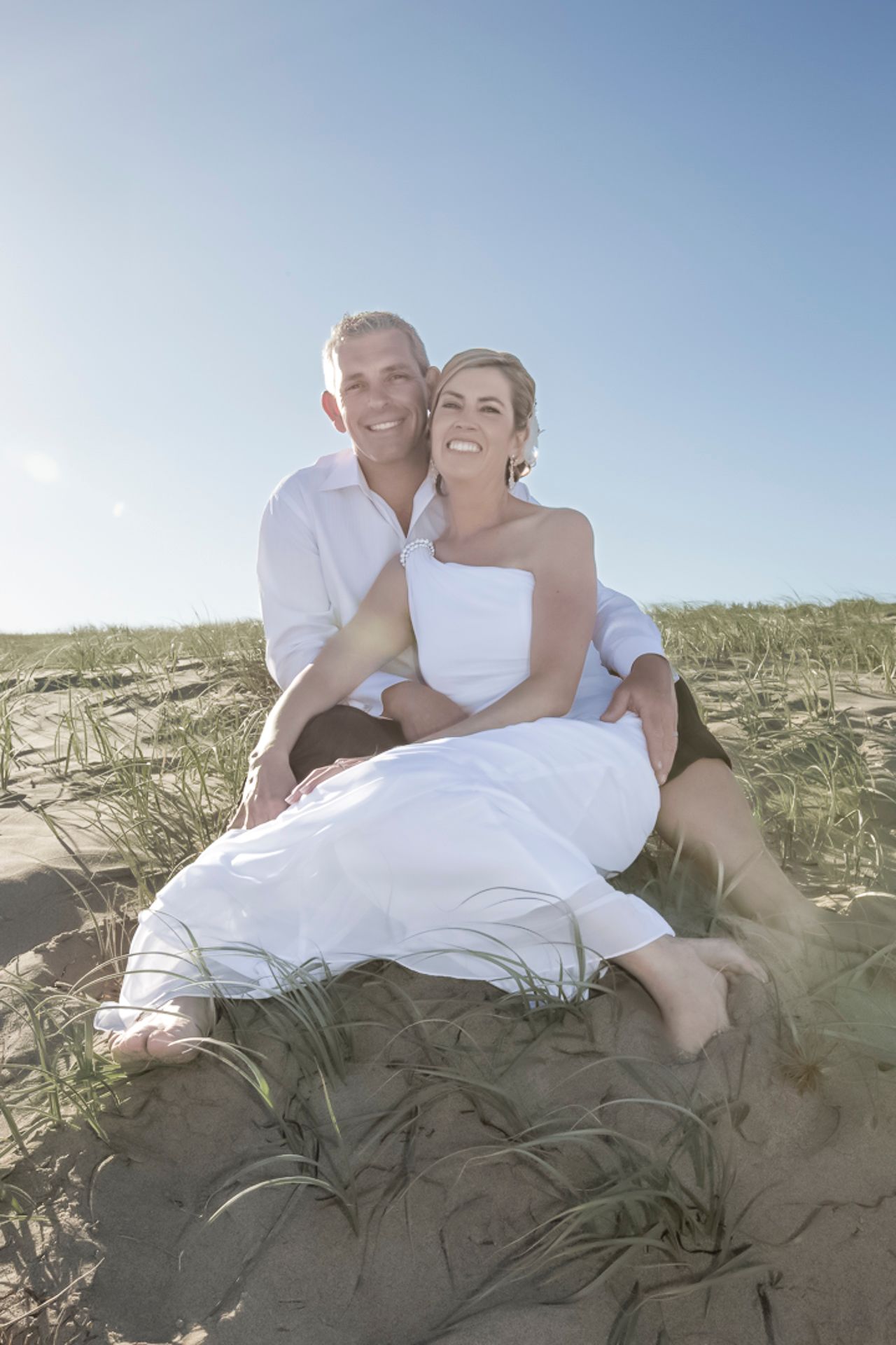 A couple in white wedding attire sits casually on a sandy beach dune under a bright blue sky.