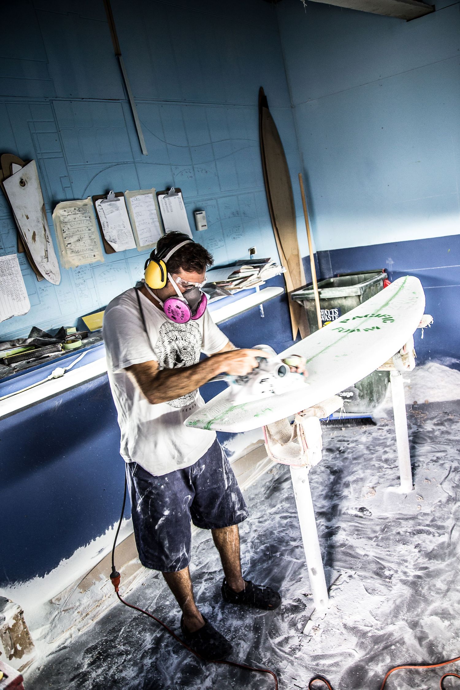 A surfboard shaper works meticulously on shaping a white surfboard in a foam-covered workshop space.
