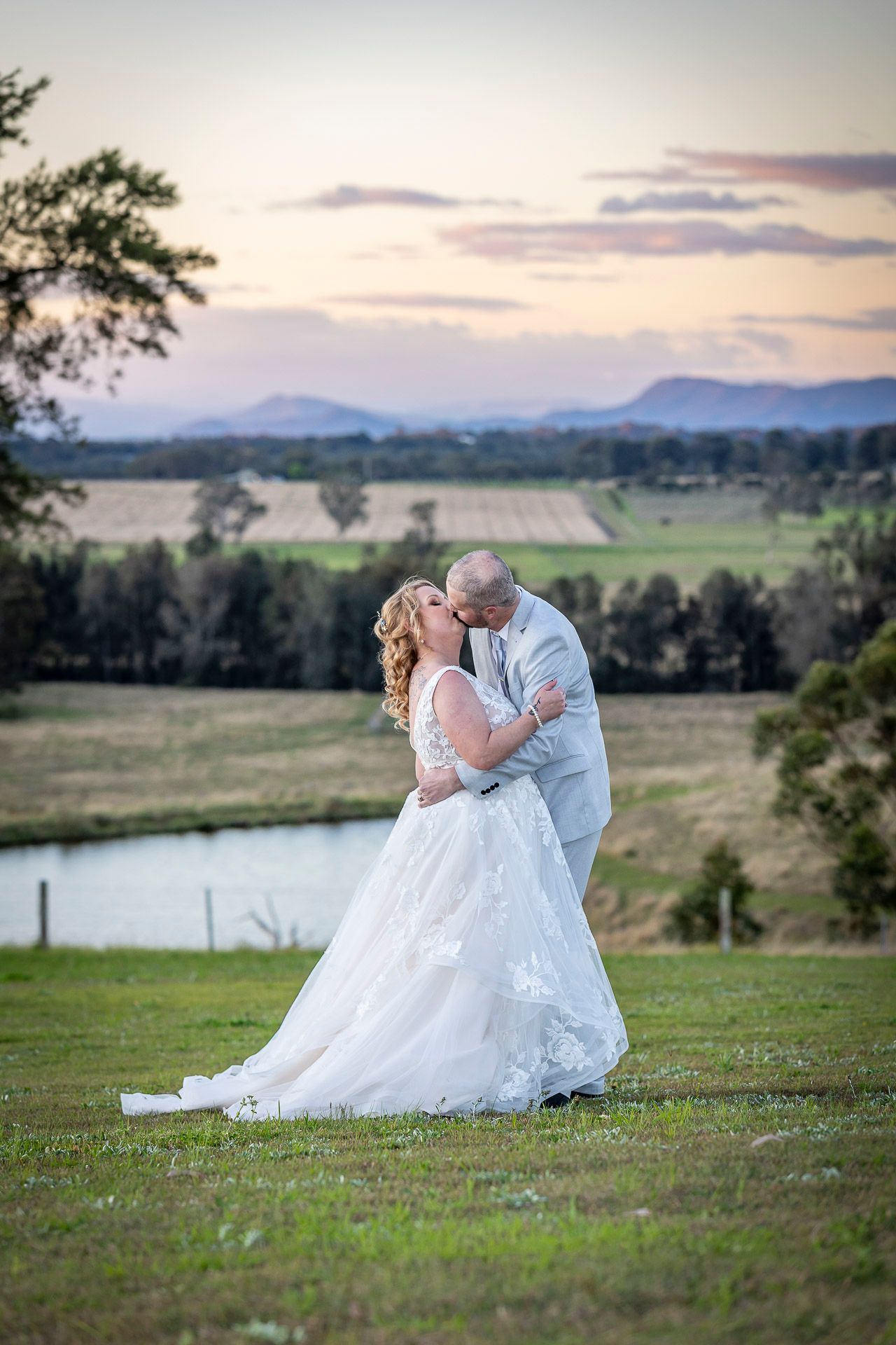 A romantic couple in wedding attire share an intimate moment on a grassy hillside overlooking a scenic landscape at sunset.