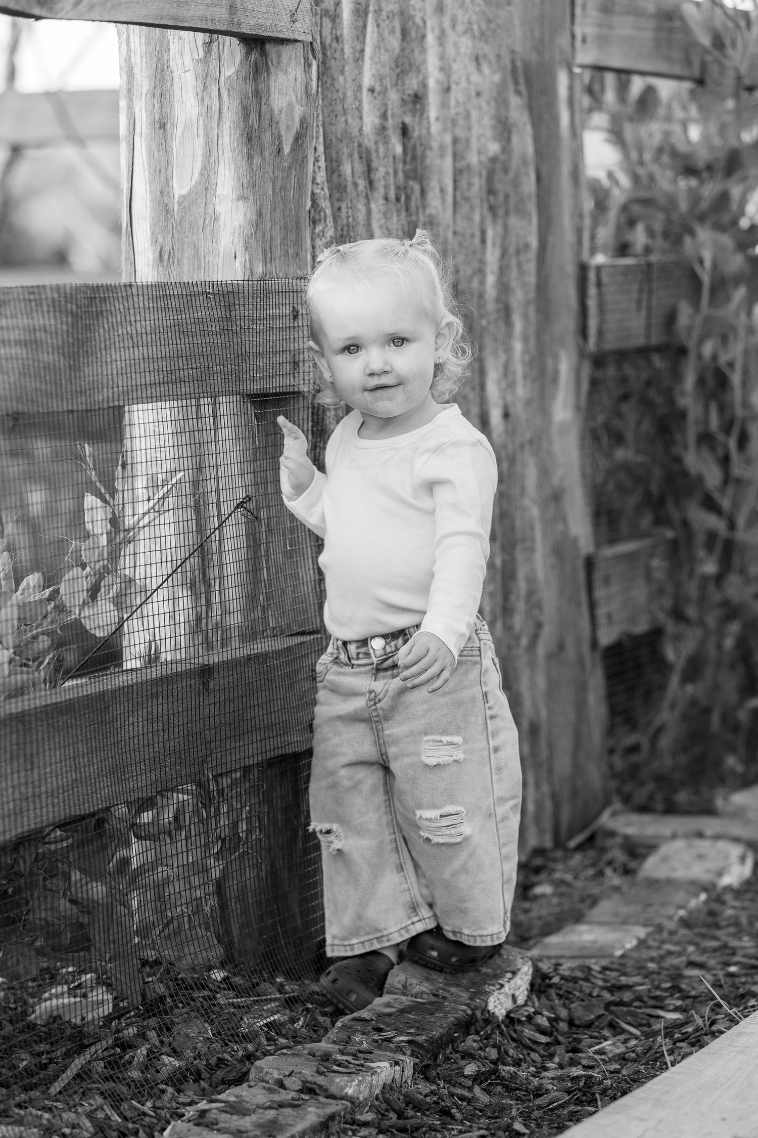 Black and white portrait of a small child standing by a wooden fence wearing jeans and a light colored shirt.